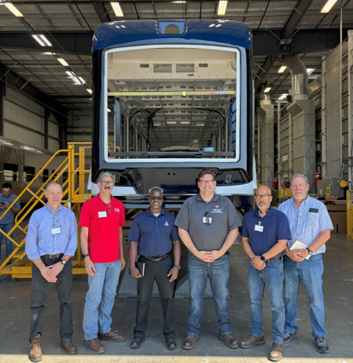 From left, Don Haradem, Michael Schipper, Dr. Floun’say Caver, Casey Blaze, Bryan Moore, and Ken Bisson stand in front of the carshell at the manufacturing facility.